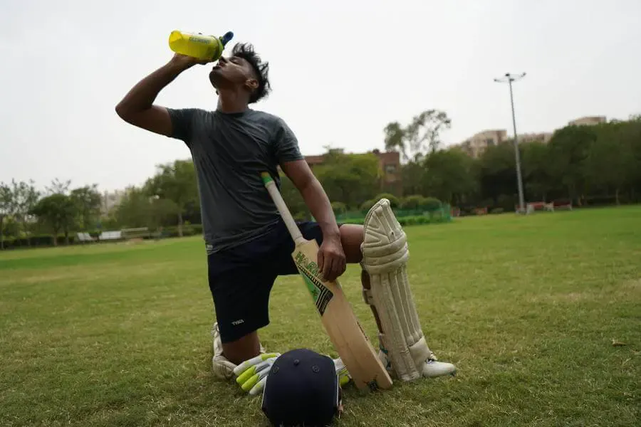 A cricket player in sports attire and leg guard kneels on one leg with his cricket bat against his torso and drinks from a yellow bottle with his head tilted back. Cricket helmet and gloves are on the ground of the field in front of him.