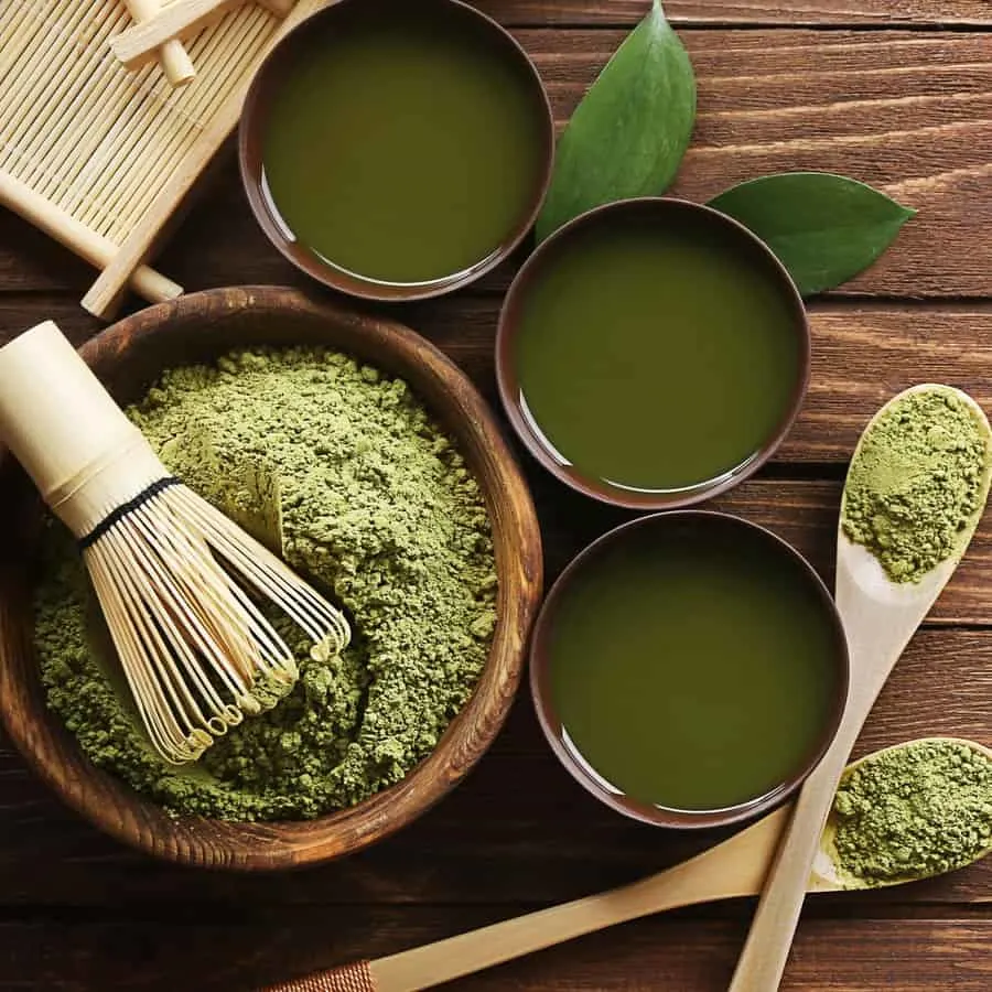 Green tea powder in a bowl along with green tea cups and spoons full of green tea.