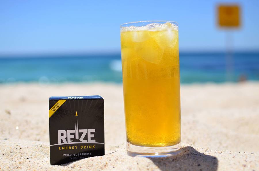 Glass of REIZE with its packet to the side on a sandy beach.