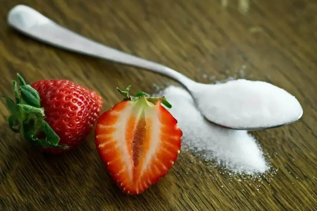 A spoon of sugar next to a couple of strawberries on a table. 