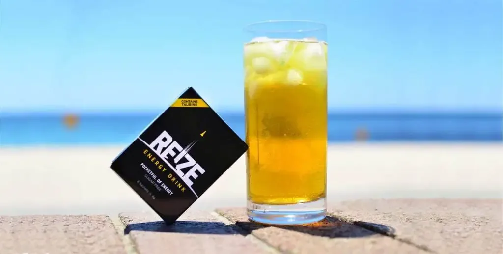 A glass of REIZE next to a sachet of REIZE on a beach. 