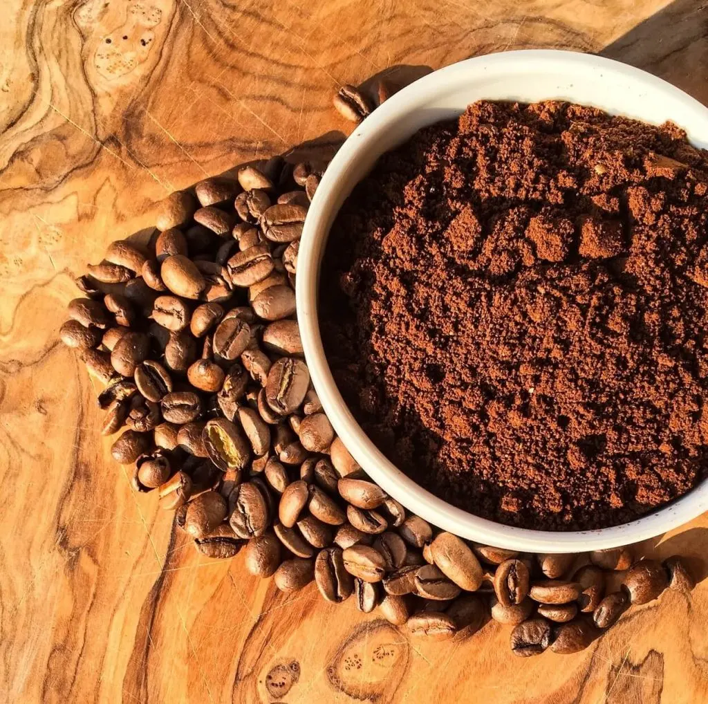 A white bowl contains caffeine powder while being surrounded by coffee beans on a table. 