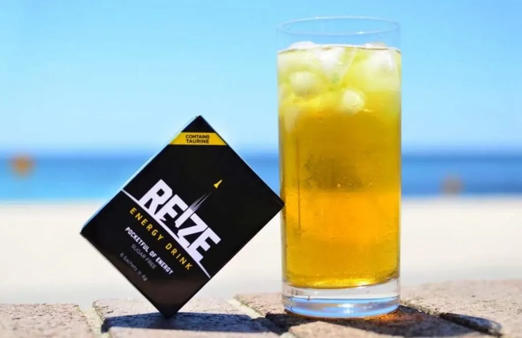 A glass of REIZE with a sachet of REIZE in front of the beach. 