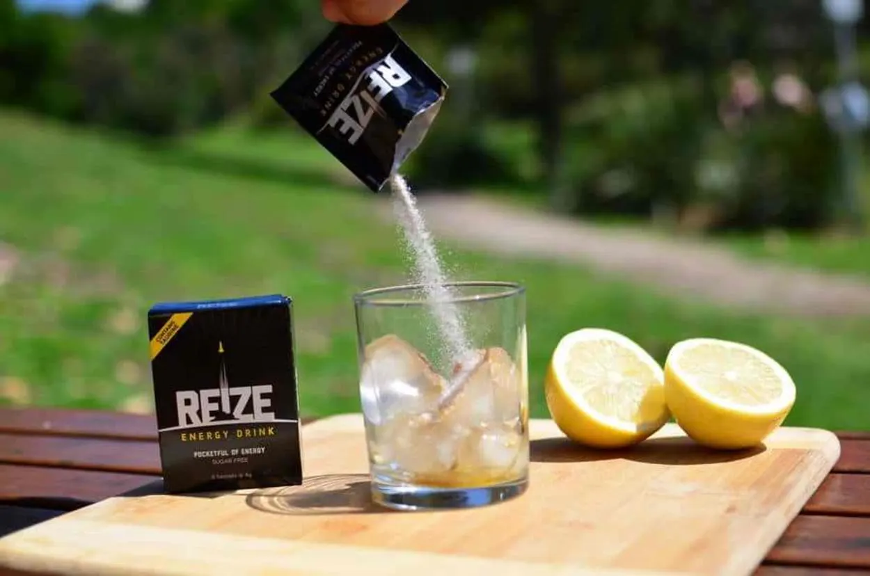 A pack of REIZE energy drink poured into a glass full of ice. A pack of REIZE energy drink and a sliced lemon can be seen in the background.